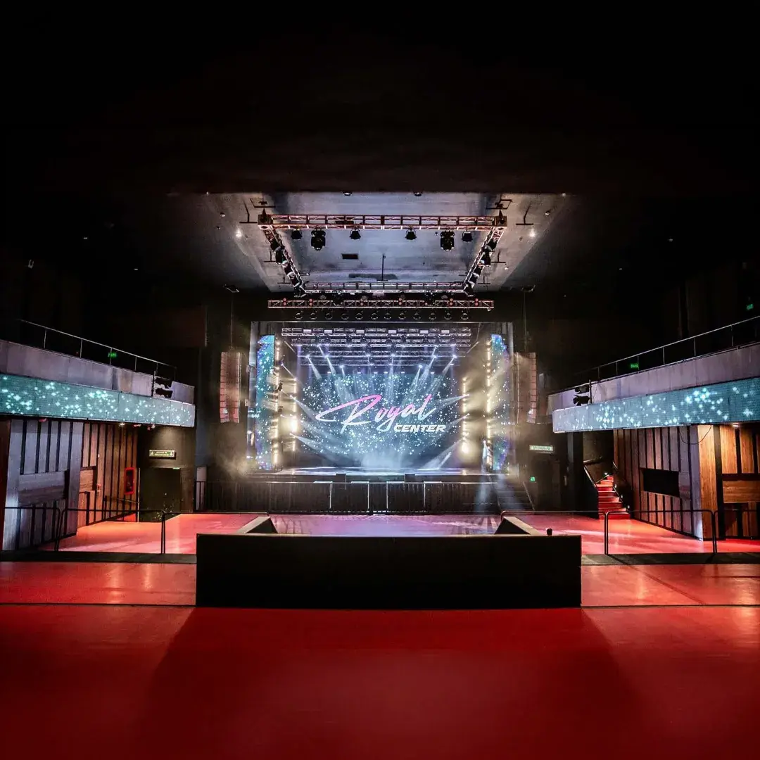 Salón del Royal Center con el escenario iluminado y la pantalla al fondo.