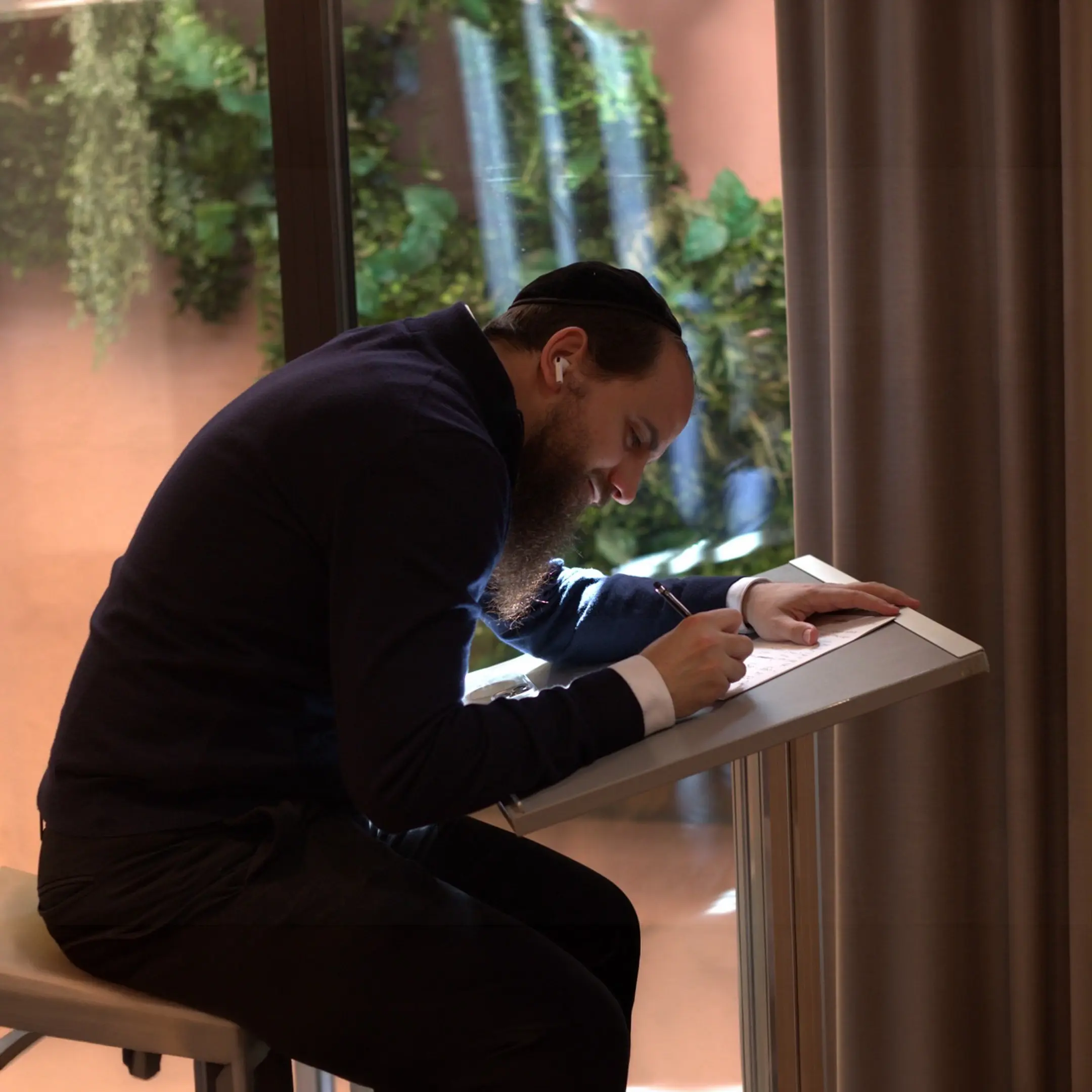 Daniel R. Chapán escribe inclinado sobre un atril, junto a una ventana con plantas.