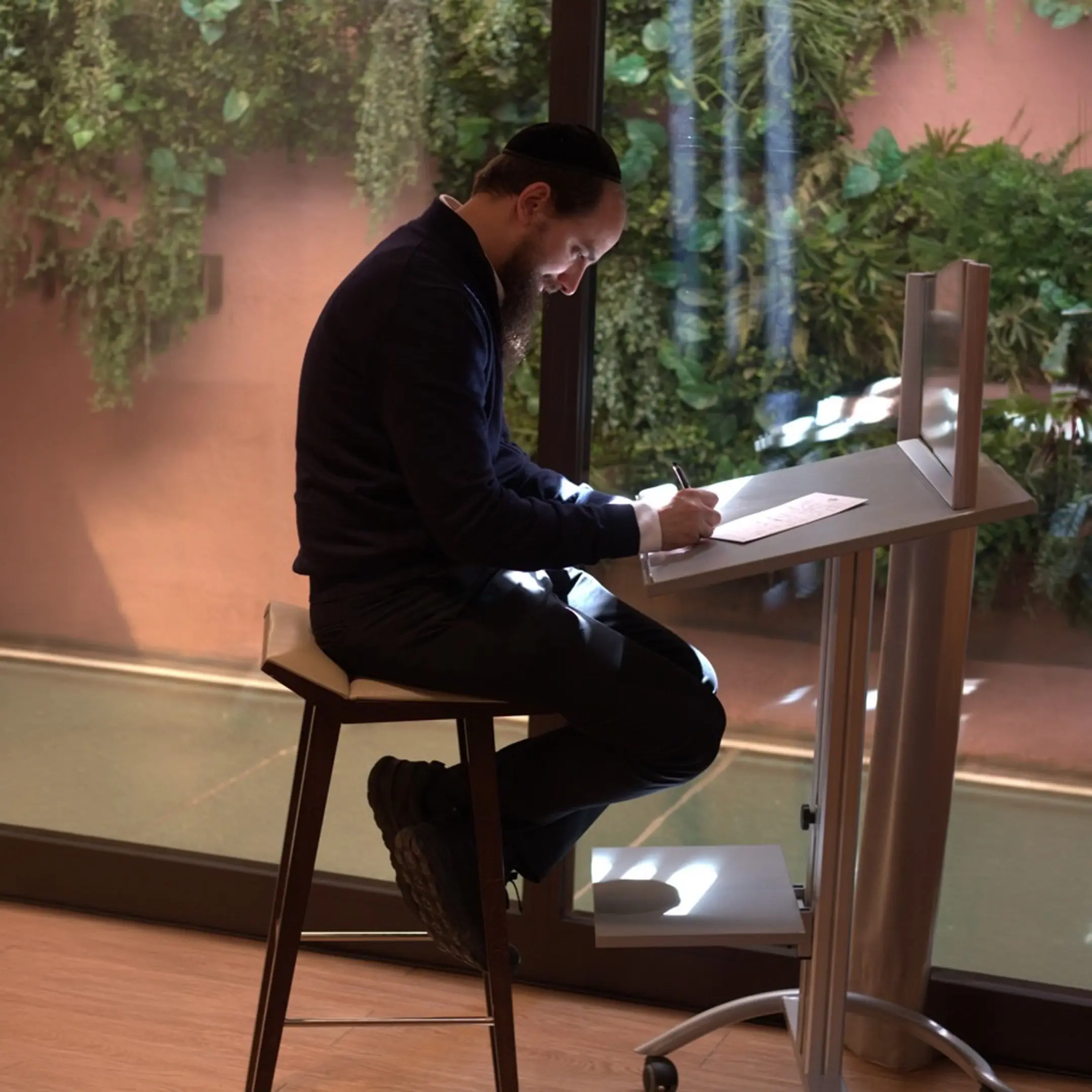 Daniel R. Chapán, sentado en un banco alto, escribe en un atril frente a un jardín vertical.